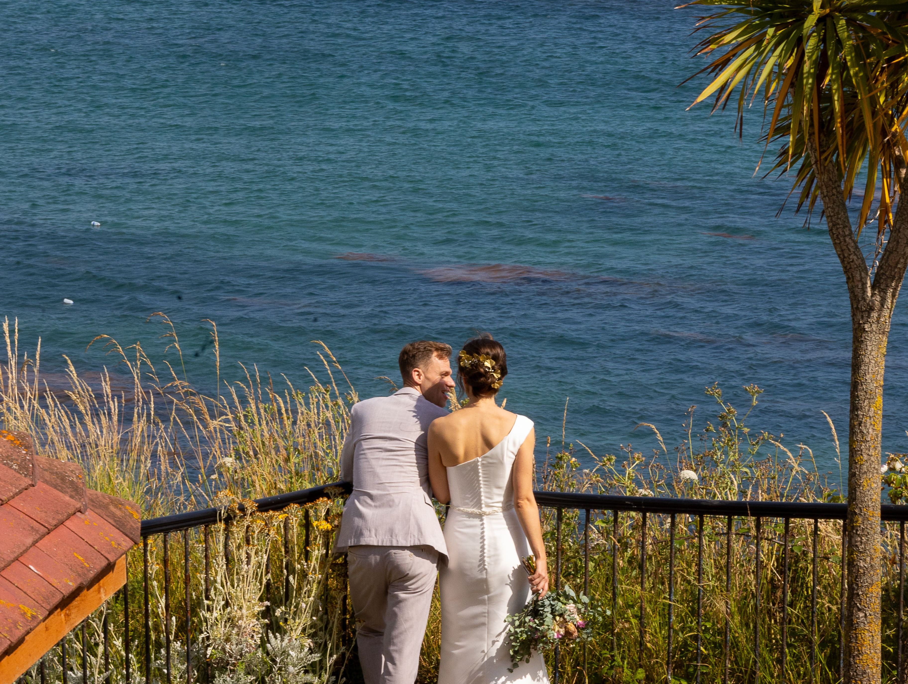 Bride and groom standing by a railing overlooking the sea with a sailboat in the distance.