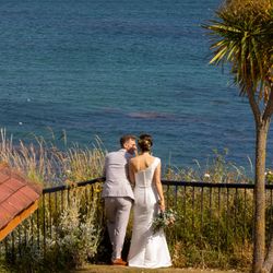 Bride and groom standing by a railing overlooking the sea with a sailboat in the distance.