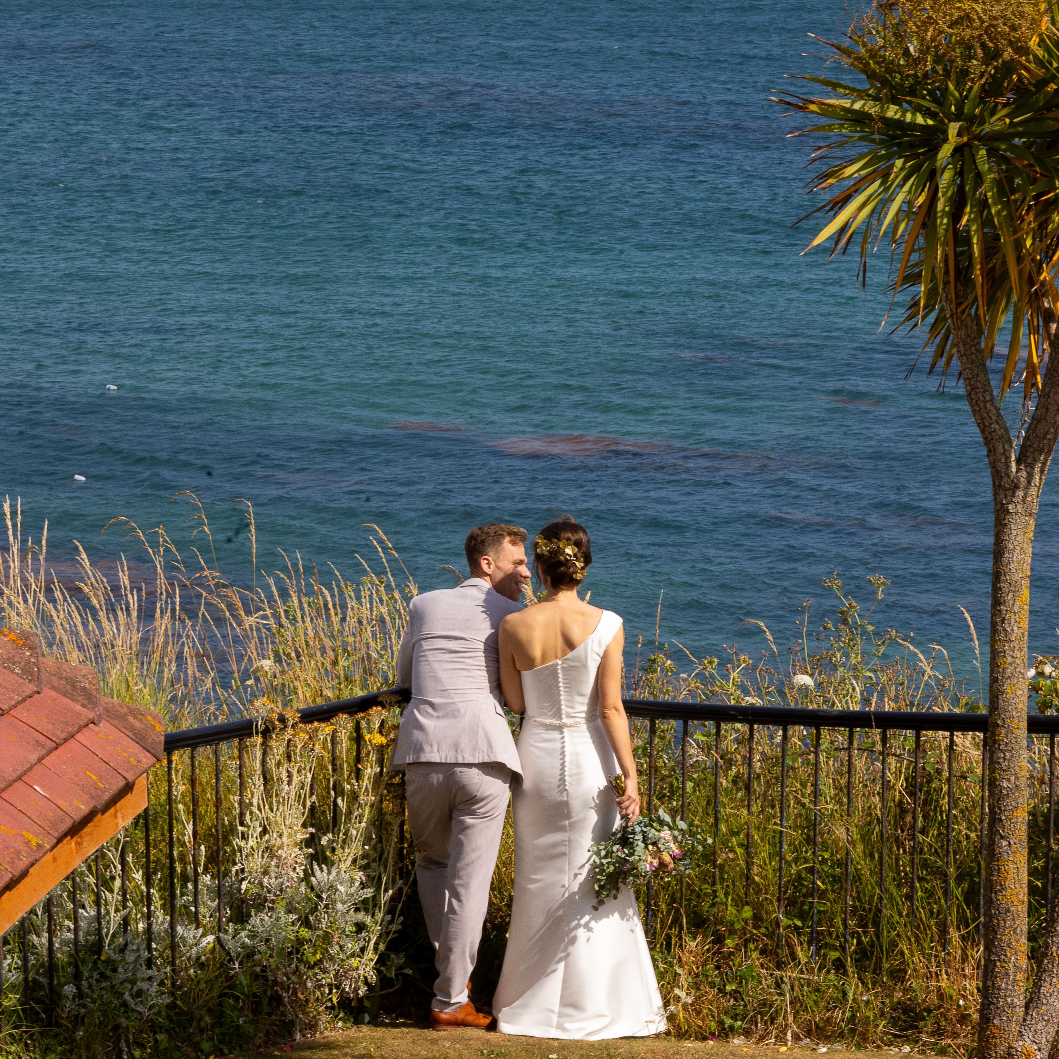 Bride and groom standing by a railing overlooking the sea with a sailboat in the distance.