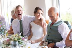 A group of people sitting at a wedding reception table, with one man laughing heartily, a bride smiling, and an older man looking down and smiling.