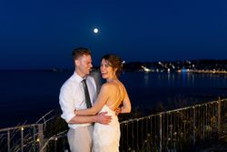 A happy couple in wedding attire embracing and smiling at night by the water with the moon and city lights in the background.
