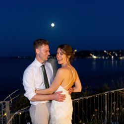 A happy couple in wedding attire embracing and smiling at night by the water with the moon and city lights in the background.