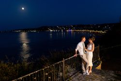 Couple standing together by a railing overlooking a calm sea at night, with city lights and a full moon reflecting on the water.