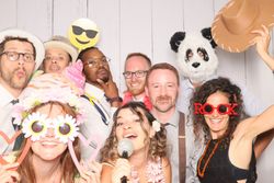 A group of people in a photo booth wearing playful costumes and props, including hats, novelty glasses, a panda mask, and holding fun signs.