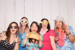 Five women in colorful outfits and fun accessories posing happily in front of a white wooden wall.