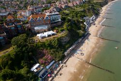 Aerial view of a seaside town with a sandy beach, colorful beach huts, and cliffs.