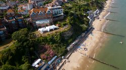Aerial view of a seaside town with a sandy beach, colorful beach huts, and cliffs.