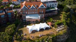 Aerial view of a large building with a red-tiled roof, a white marquee tent on the lawn, and garden terraces on a cliff edge.