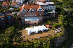 Aerial view of a large building with a red-tiled roof, a white marquee tent on the lawn, and garden terraces on a cliff edge.