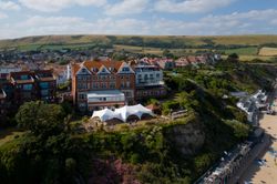 Aerial view of a seaside hotel perched on a cliff above a beach, with a large white tent in the garden and a town and rolling hills in the background.