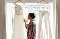 Woman smiling at a hanging wedding dress and bridal pantsuit by a window
