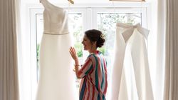 Woman smiling at a hanging wedding dress and bridal pantsuit by a window