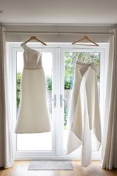 Two elegant white bridal outfits hanging on hangers in front of a glass door.