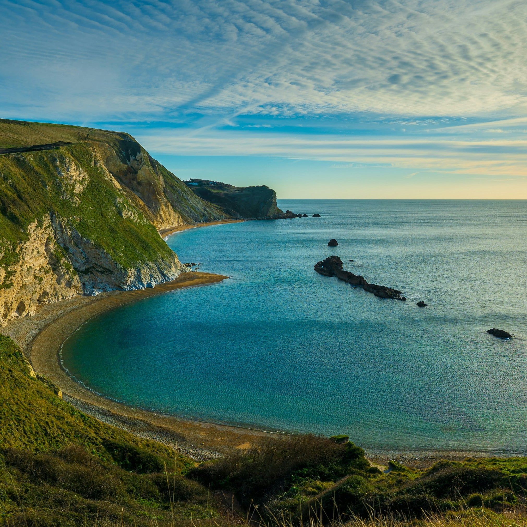 Curved coastline with green cliffs, clear blue water, and a rock formation extending into the sea under a partly cloudy sky.