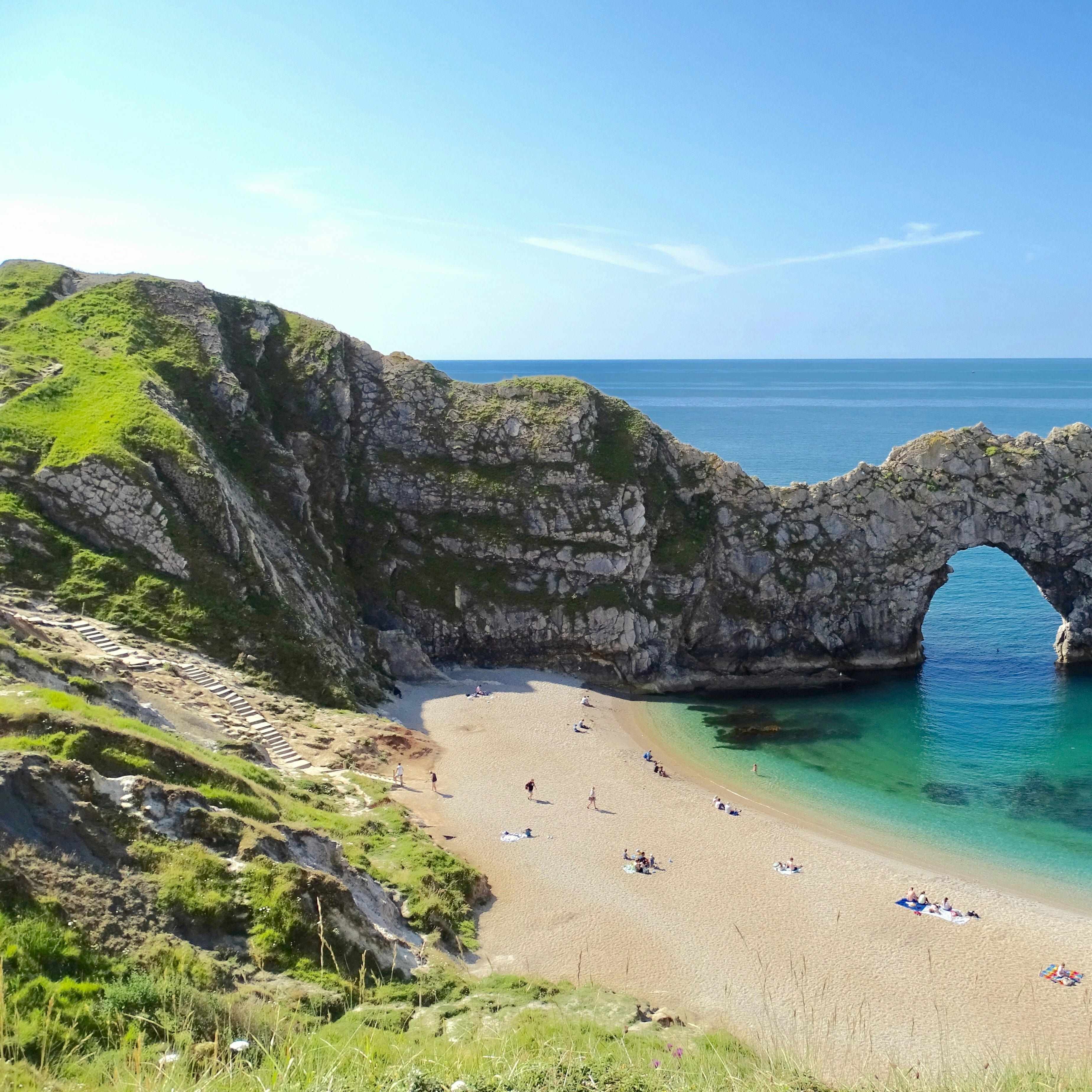 Natural limestone arch overlooking a sandy beach and clear blue sea with people relaxing on the shore