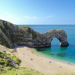 Natural limestone arch overlooking a sandy beach and clear blue sea with people relaxing on the shore