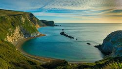 A scenic coastal bay with steep cliffs, a pebbled beach, and rocky outcrops in the water under a partly cloudy sky at sunset.