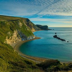 A scenic coastal bay with steep cliffs, a pebbled beach, and rocky outcrops in the water under a partly cloudy sky at sunset.