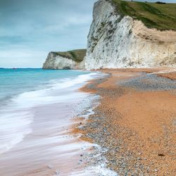 Steep white chalk cliffs beside a pebbled beach and turquoise sea under a cloudy sky