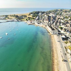 Aerial view of a seaside town with a sandy beach, clear turquoise water, boats, and a pier extending into the sea.