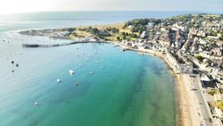 Aerial view of a seaside town with a sandy beach, clear turquoise water, boats, and a pier extending into the sea.