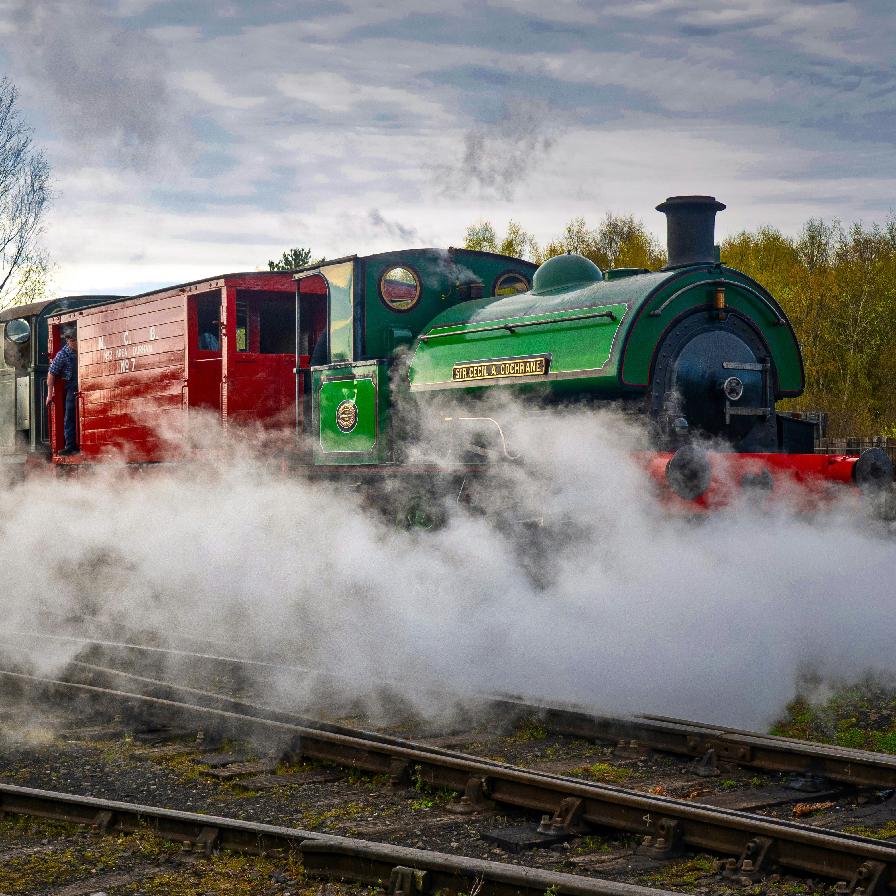 Vintage green steam locomotive emitting steam on railway tracks with a red carriage attached.