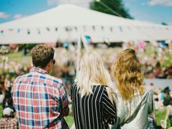 Three people standing together at an outdoor festival, viewed from behind, with a large tent and a crowd in the background.