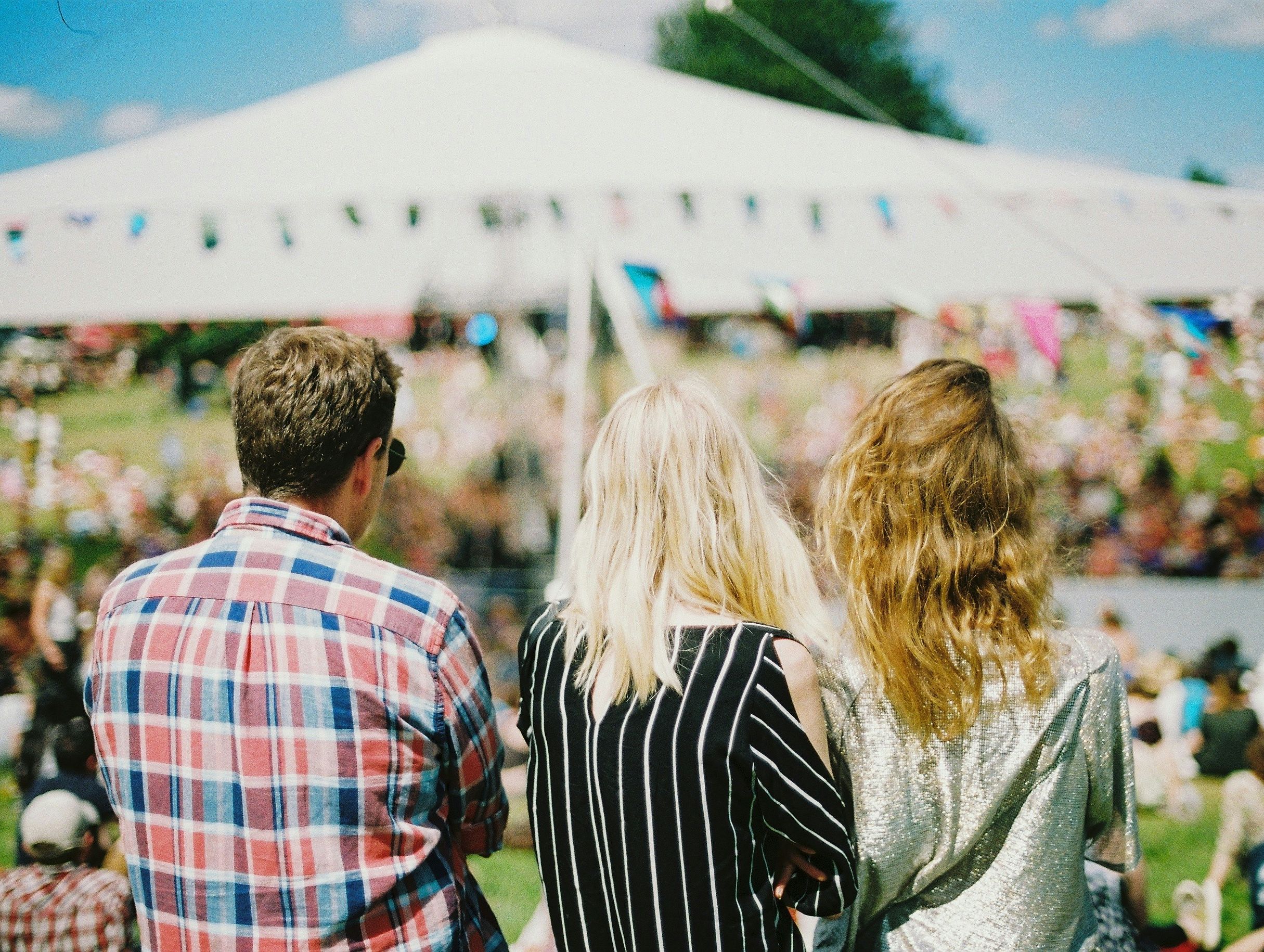 Three people standing together at an outdoor festival, viewed from behind, with a large tent and a crowd in the background.