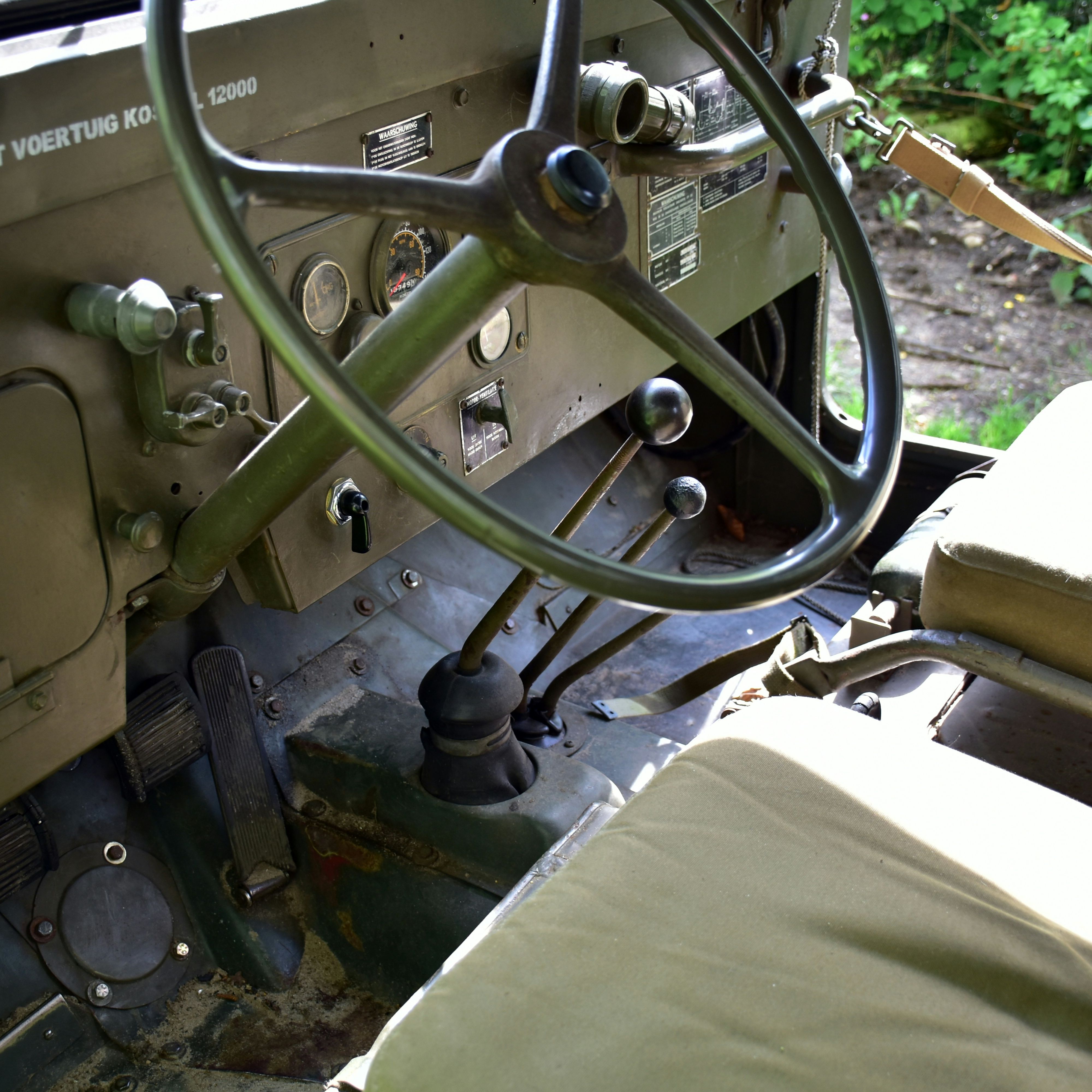 Interior view of a vintage military vehicle showing the steering wheel, dashboard, gear levers, pedals, and olive green seats.