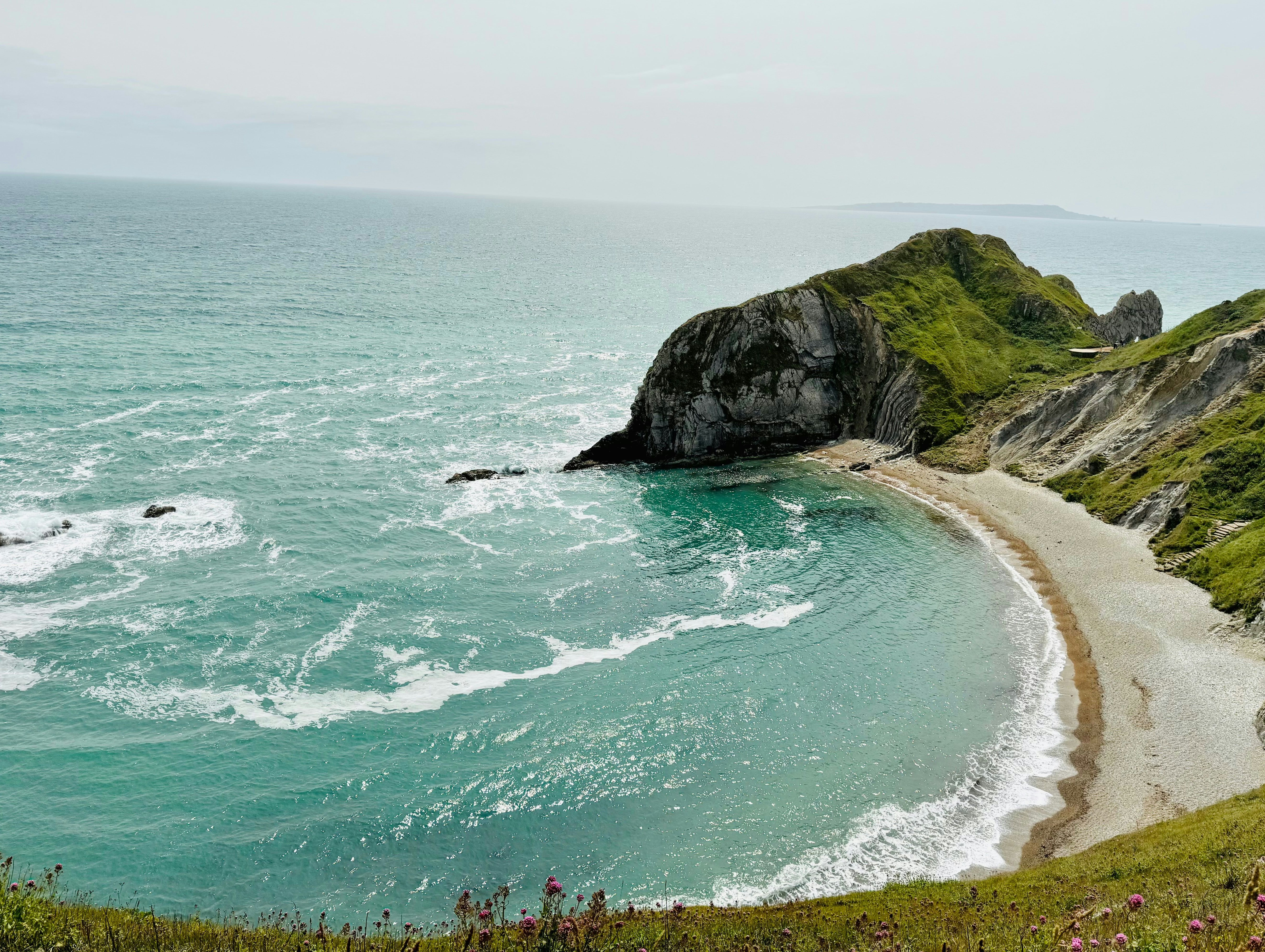 Scenic coastal view with turquoise ocean waves meeting a sandy, crescent-shaped beach bordered by green cliffs.