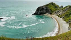 Scenic coastal view with turquoise ocean waves meeting a sandy, crescent-shaped beach bordered by green cliffs.