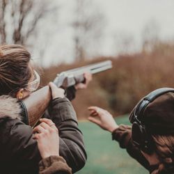 Two people outdoors, one aiming a shotgun while the other provides instruction, both wearing safety gear
