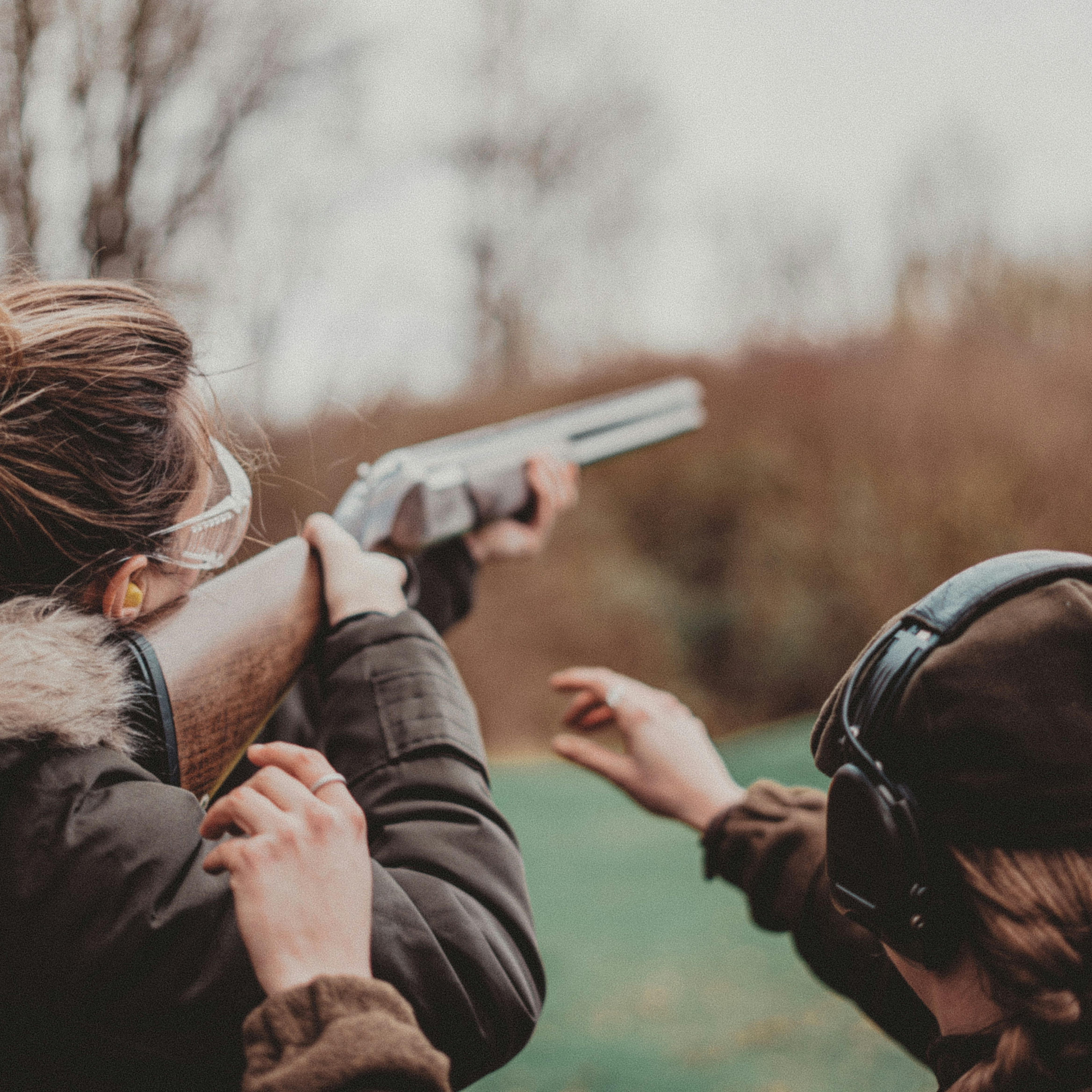 Two people outdoors, one aiming a shotgun while the other provides instruction, both wearing safety gear