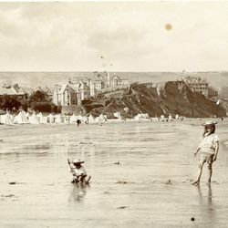 Vintage photograph of children playing on a beach with large hats, with tents and Victorian-style buildings in the background.