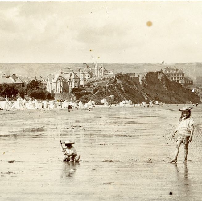 Vintage photograph of children playing on a beach with large hats, with tents and Victorian-style buildings in the background.