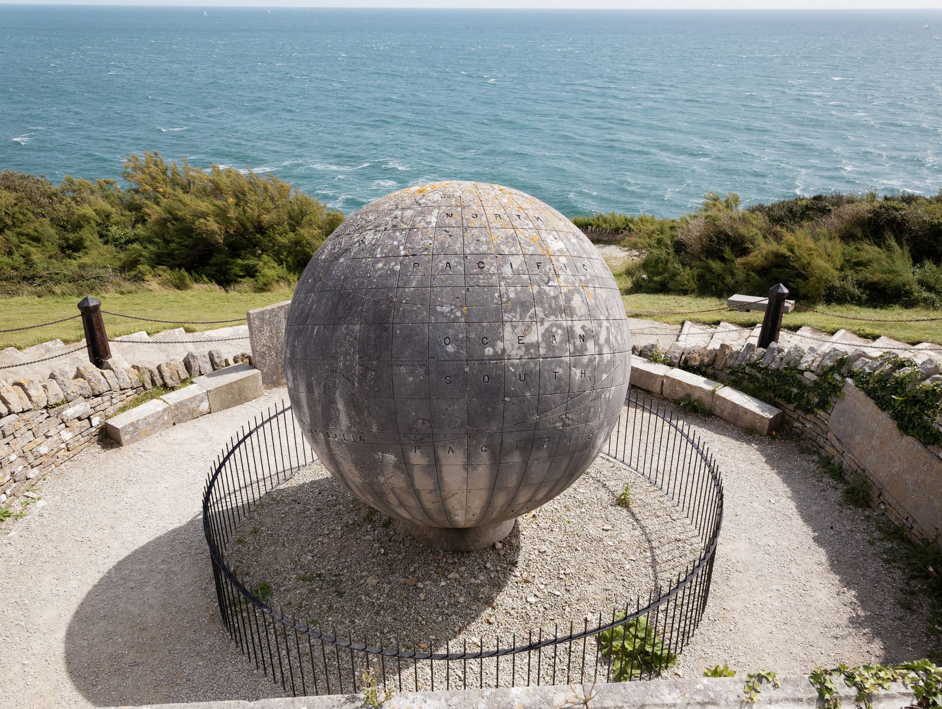A large stone globe monument overlooking the sea, surrounded by a circular stone wall and greenery.