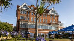 A large red-brick building with white gables and many windows, likely a hotel, surrounded by a garden with picnic benches, blue umbrellas, palm trees, and purple flowers in the foreground.