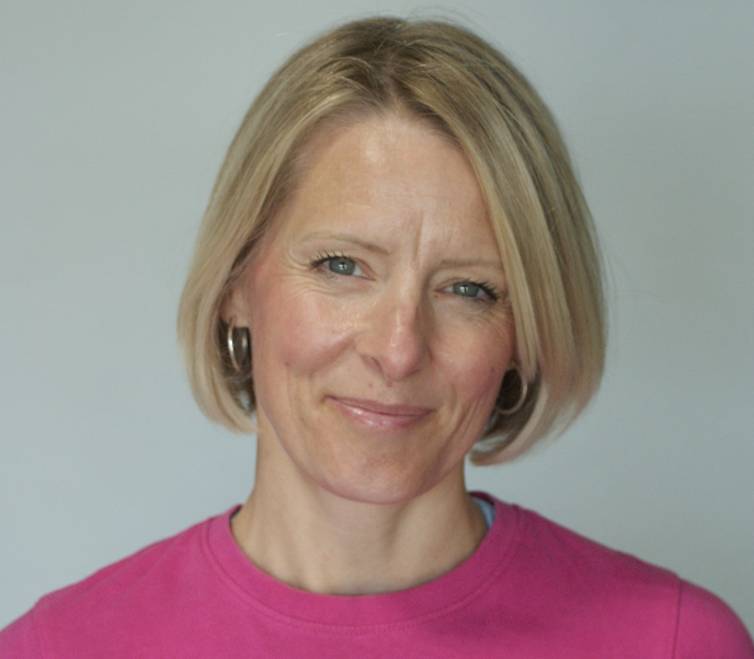 Woman with short blonde hair wearing a pink 'Su Evans Fitness' t-shirt, smiling at the camera