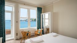 Bright bedroom with a sea view through large windows, featuring two wicker chairs and a bed with white linens.