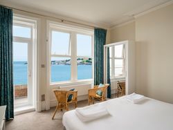 Bright bedroom with a sea view through large windows, featuring two wicker chairs and a bed with white linens.