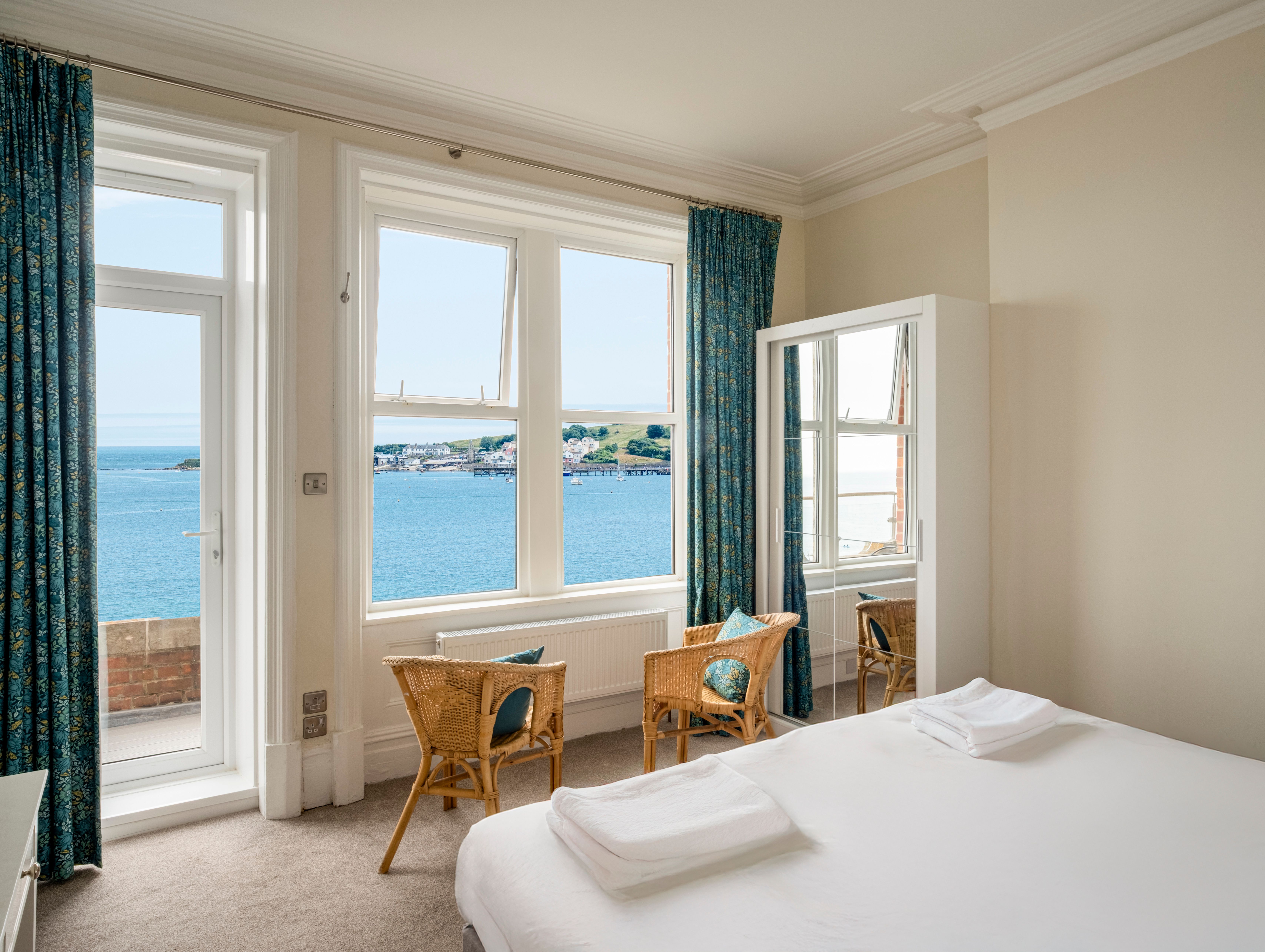 Bright bedroom with a sea view through large windows, featuring two wicker chairs and a bed with white linens.