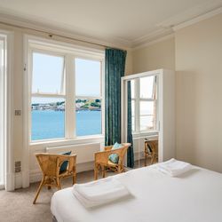 Bright bedroom with a sea view through large windows, featuring two wicker chairs and a bed with white linens.