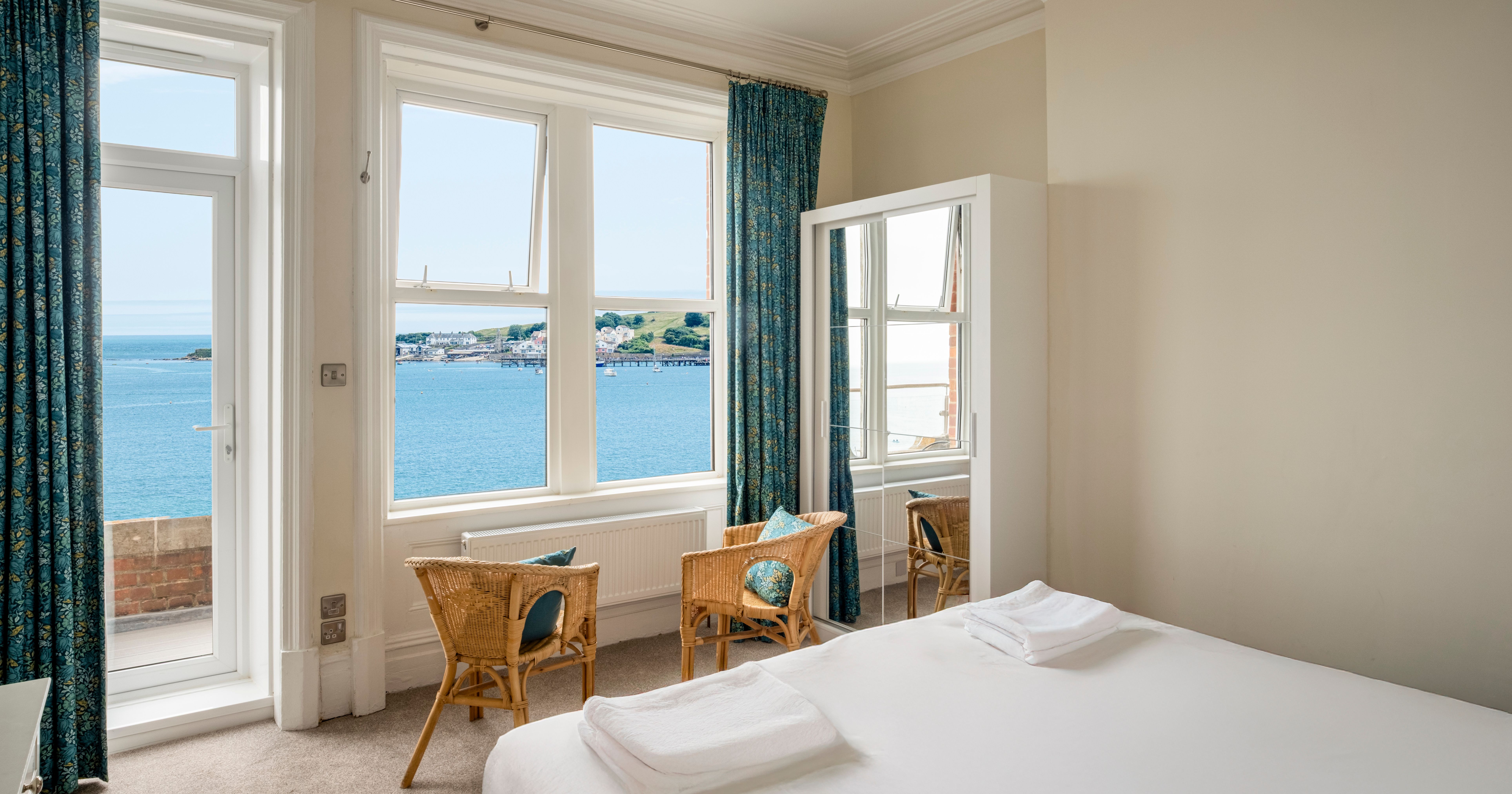 Bright bedroom with a sea view through large windows, featuring two wicker chairs and a bed with white linens.