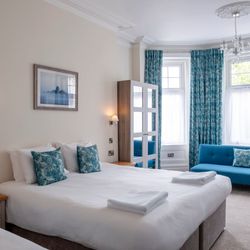 Bright hotel bedroom with double bed, blue throw pillows, blue sofa, and large windows with patterned blue curtains