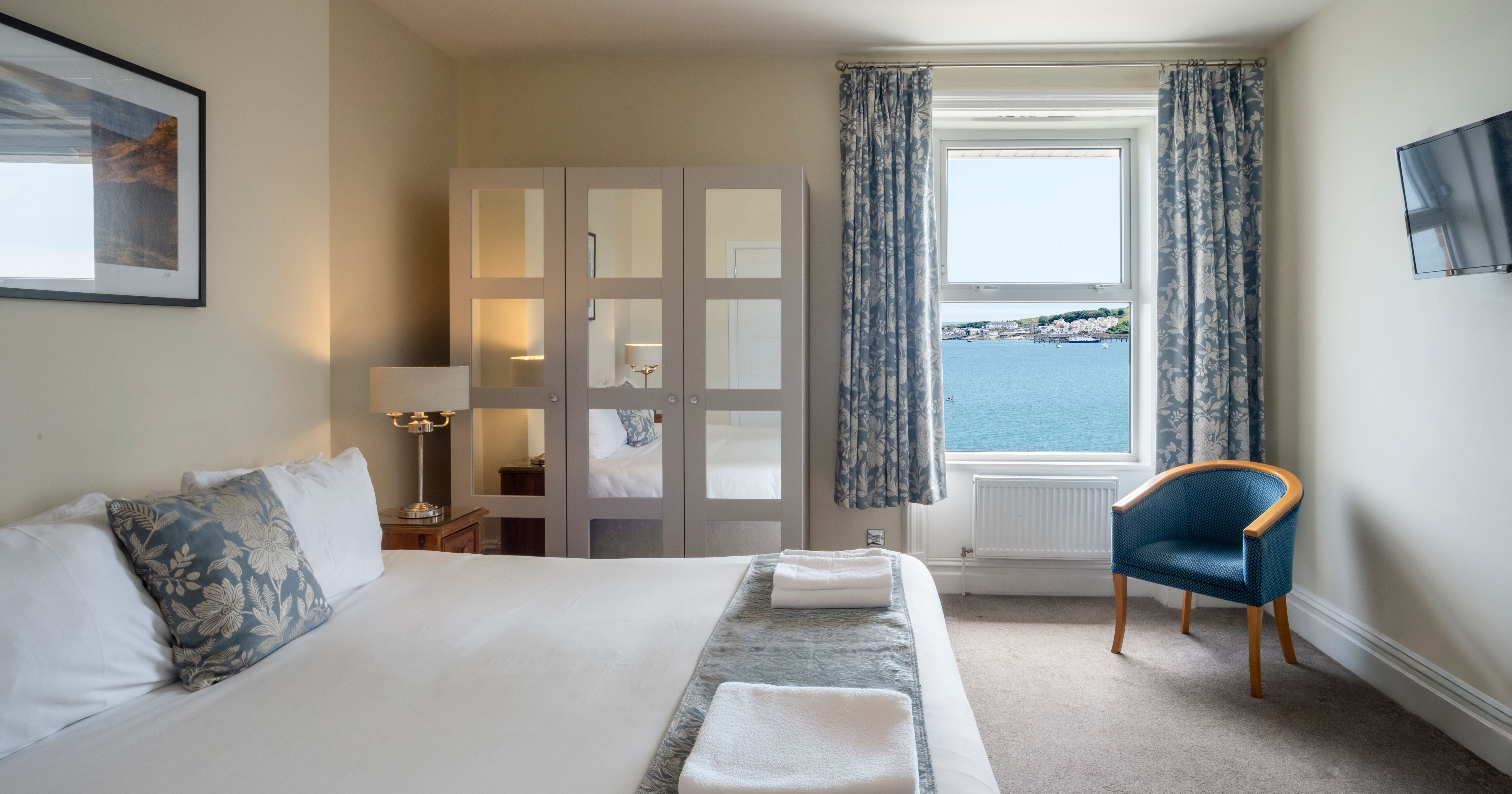 Hotel room with a large bed, mirrored wardrobe, blue floral curtains, and a window with a sea view.