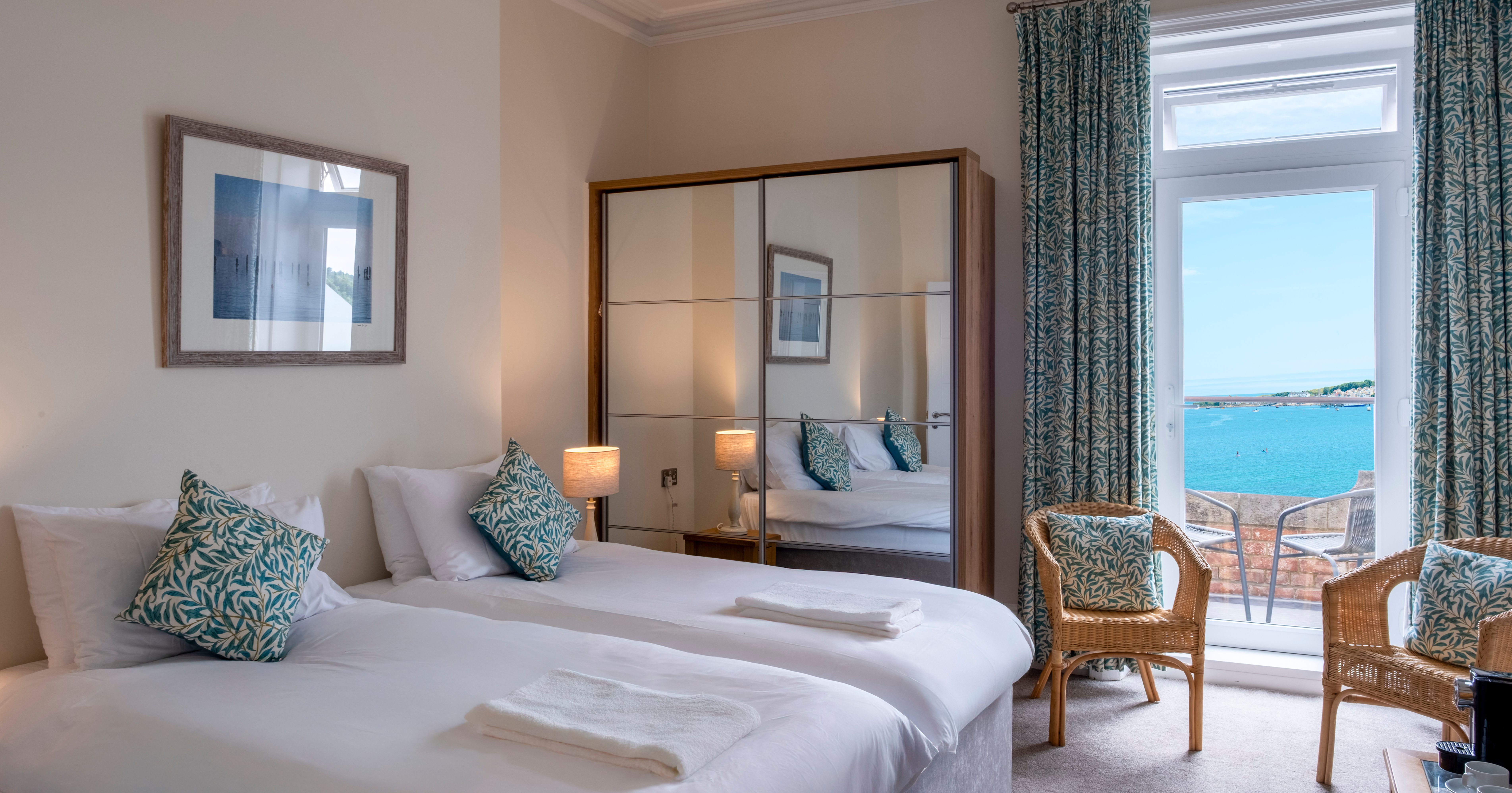 A hotel room with twin beds, a mirrored wardrobe, wicker chairs, and a view of the sea through French doors.