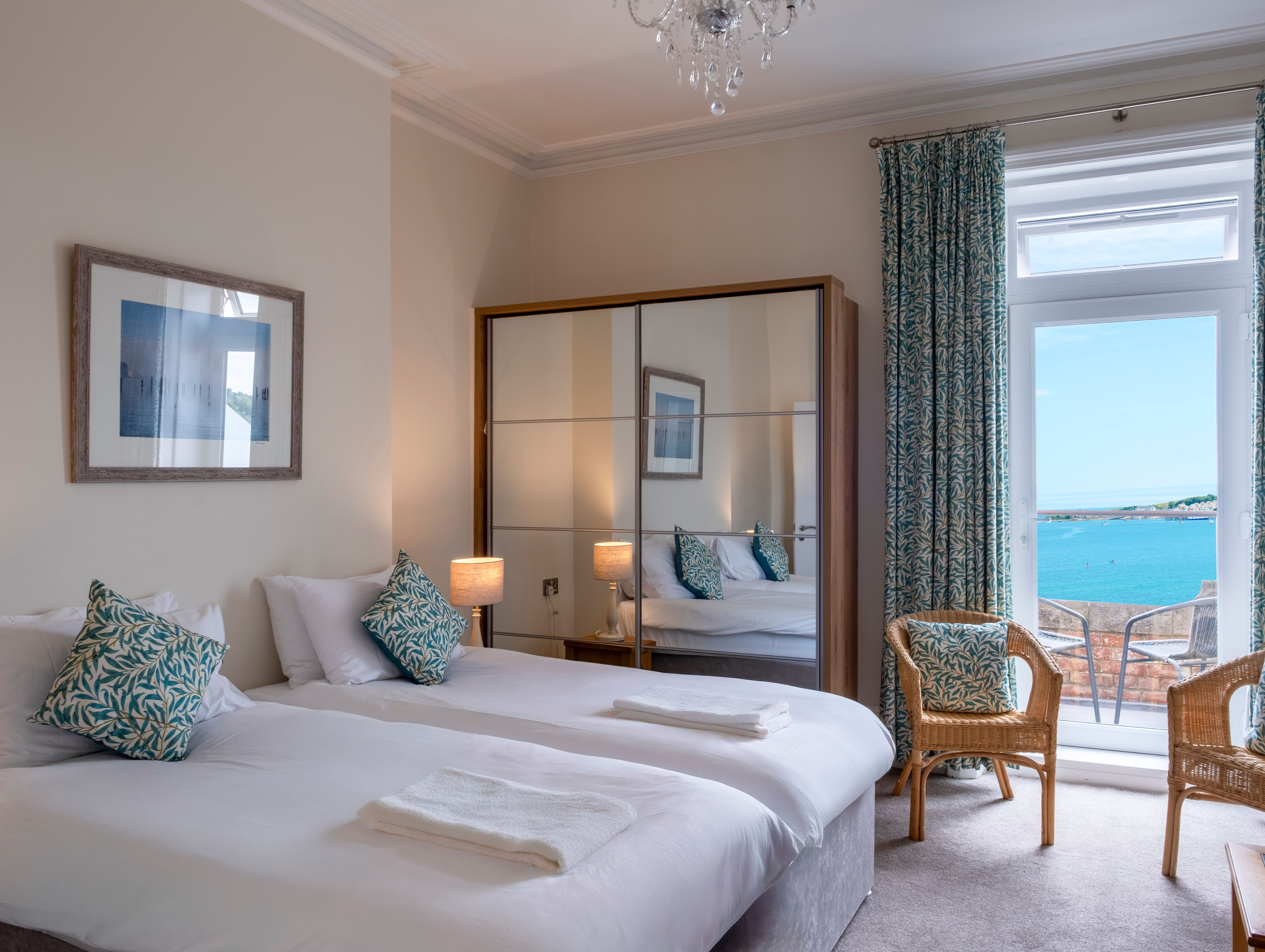 A hotel room with twin beds, a mirrored wardrobe, wicker chairs, and a view of the sea through French doors.
