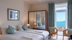 A hotel room with twin beds, a mirrored wardrobe, wicker chairs, and a view of the sea through French doors.