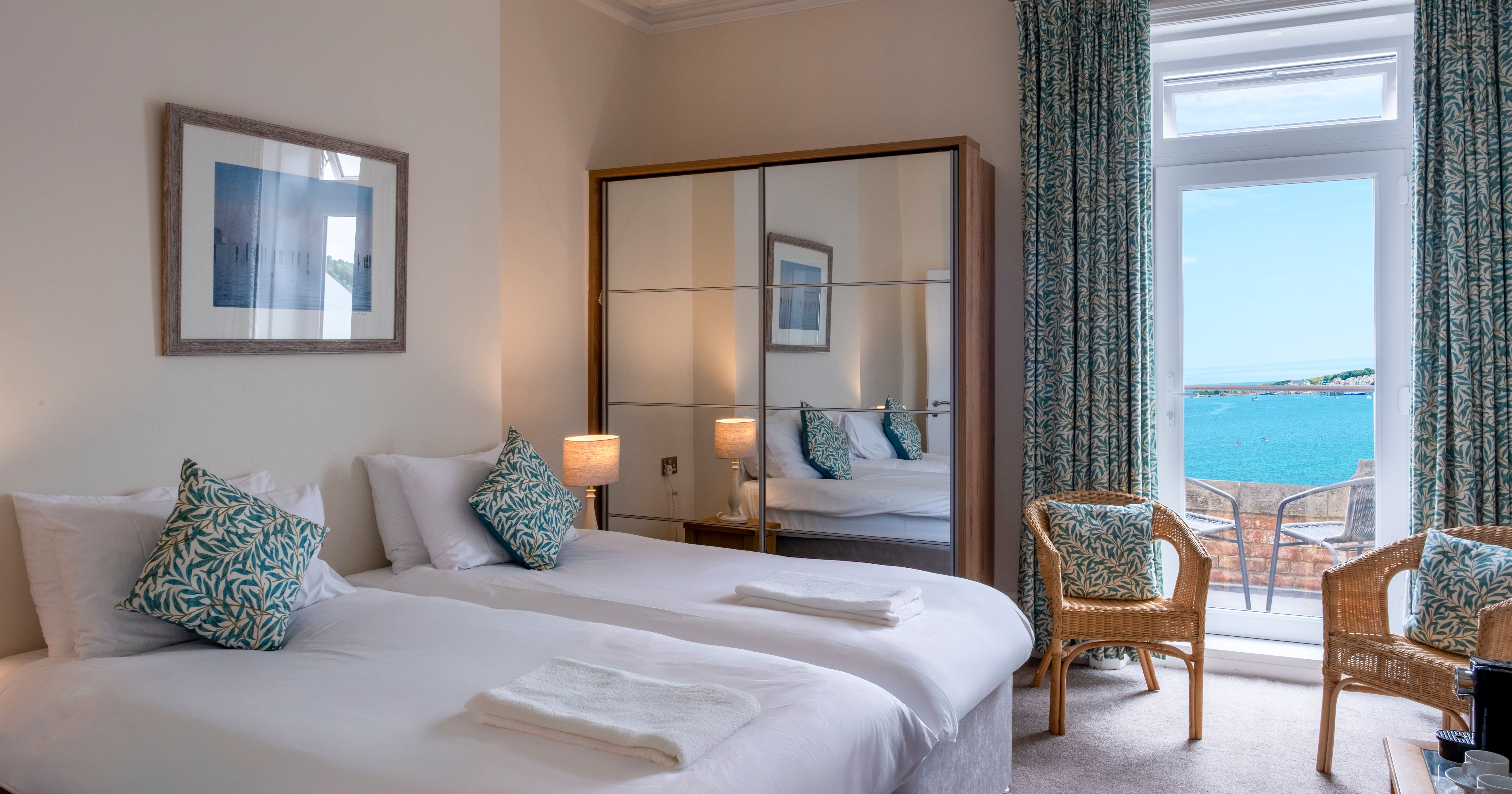 A hotel room with twin beds, a mirrored wardrobe, wicker chairs, and a view of the sea through French doors.