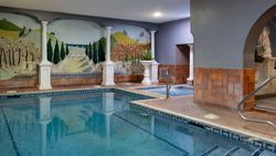 Indoor swimming pool with mosaic tiles, a hot tub area, and decorative mural on the wall featuring trees, columns, and landscape art.
